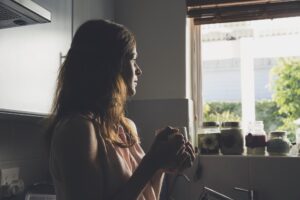 Woman at a kitchen window holding a mug, looking thoughtful — illustrating the daily work of staying positive